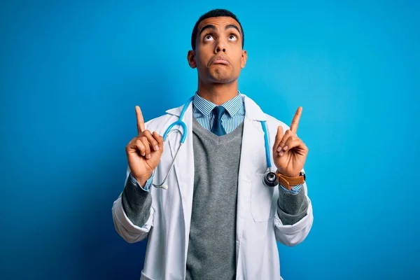 英俊的非洲医生 身穿外套 带着听诊器 头戴蓝色背景的美国医生 带着悲伤和不安的表情 用手指指示方向 不快乐和沮丧 — 图库照片