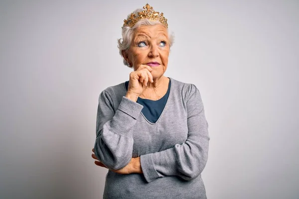 Senior beautiful grey-haired woman wearing golden queen crown over white background with hand on chin thinking about question, pensive expression. Smiling with thoughtful face. Doubt concept.