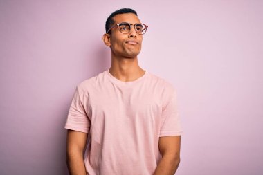 Yakışıklı Afro-Amerikalı adam pembe arka planda günlük tişört ve gözlük takıyor. Gülümsüyor ve başka tarafa bakıyor..