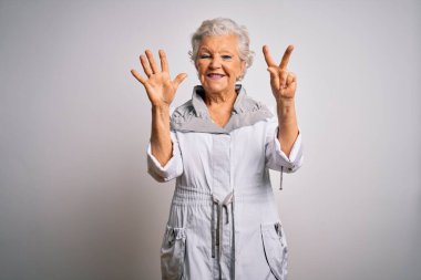 Son sınıf güzeli, gri saçlı, sıradan bir ceket giyen, beyaz arka planda duran ve yedinci parmaklarını göstererek gülümseyen, kendinden emin ve mutlu bir kadın..