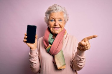 Son sınıf güzeli gri saçlı kadın elinde akıllı telefonuyla pembe arka planda ekranı gösteriyor.