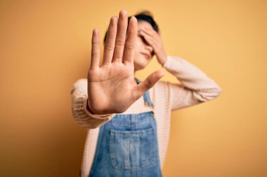 Genç, güzel Asyalı bir kız izole edilmiş sarı arka plan üzerine günlük kot pantolon giyiyor gözleri ellerle örtüyor ve üzgün ve korku ifadeleriyle dur hareketi yapıyor. Utanç verici ve olumsuz bir kavram..