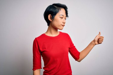 Genç, güzel Asyalı bir kız, günlük kırmızı tişört giyiyor, beyaz arka planda dikiliyor. Gururlu, gülümseyen, baş parmak kaldıran bir jest yapıyor.