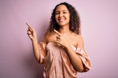 Kıvırcık saçlı, rahat tişörtlü, pembe arka planda duran, gülümseyen ve kameraya bakan genç ve güzel bir kadın..