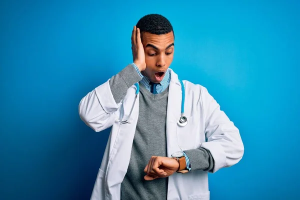 英俊的非洲裔美国医生 身穿外套 带着听诊器 背景是蓝色的 看着钟表时间 忧心忡忡 害怕迟到 — 图库照片