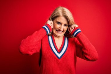 Orta yaşlı, güzel, sarışın bir kadın izole edilmiş kırmızı arka plan üzerine gündelik süveter giyip yüksek sesli müzik sesi için parmaklarıyla kulaklarını kapatıyor. Sağır kavramı.