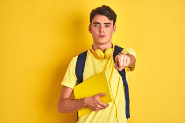 Kulaklık ve sırt çantası takan genç bir çocuk izole edilmiş arka plan üzerine kitap okuyor. Kamerayı işaret ediyor ve sen, el işareti, ön taraftan olumlu ve kendinden emin bir hareket.