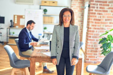 Orta yaşlı, güzel, ceketli ve gözlüklü bir iş kadını mutlu ve havalı bir gülümsemeyle ofiste dikiliyor. Şanslı kişi..