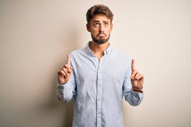 Beyaz arka plan üzerinde duran çizgili gömlekli genç yakışıklı adam üzgün ve üzgün görünüyor, parmaklarıyla yönü gösteriyor, mutsuz ve depresif..