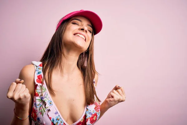 若い美しいブルネットの女性はビキニを着て休暇中にピンクの背景に帽子をかぶって 腕を上げて 笑顔で 成功のために叫んでいる勝者のジェスチャーをすることは非常に幸せで興奮しています お祝いのコンセプト — ストック写真
