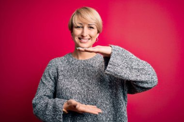 Modern kısa saçlı genç bir kadın pembe arka planda gelişigüzel süveter giyiyor. Elleri büyük ve büyük boy işareti, ölçü sembolü gösteriyor. Kameraya bakıp gülümsüyor. Ölçüm konsepti.