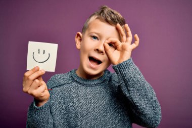 Küçük beyaz çocuk gülen suratını kâğıt bir nota gösteriyor. Mutlu yüzlü, gülümseyen bir mesaj. Tamam.