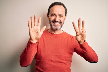 Orta yaşlı, turuncu süveter giyen, izole edilmiş beyaz arka plan üzerinde duran, 8 numaralı parmaklarıyla işaret eden, kendinden emin ve mutlu gülümseyen, orta yaşlı bir adam..