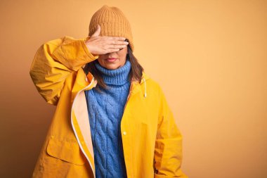 Sarı yağmurluk ve kış şapkası takan orta yaşlı bir kadın izole edilmiş arka planda gülümsüyor ve elleri yüzünde şaşkınlıktan gözleri kapayarak gülüyor. Kör kavram.