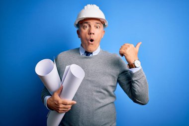 Orta yaşlı, yakışıklı, gri saçlı, kasklı bir mimar elinde planlar, elleri yana dönük, ağzı açık, ağzı açık, şaşırmış bir şekilde işaret ediyor..