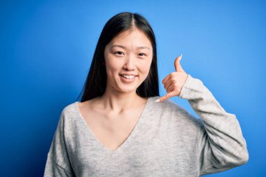 Genç ve güzel Asyalı bir kadın mavi izole edilmiş arka planda dikilmiş telefonda el ve parmaklarıyla telefonda konuşur gibi gülümsüyor. İletişim kavramları.