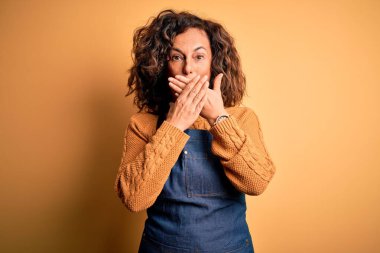 Orta yaşlı, güzel fırıncı kadın önlük giyip izole edilmiş sarı arka planda dikiliyor. Ağzını yanlışlıkla ellerle kapatıyor. Gizli konsept.