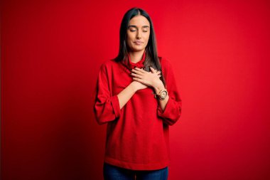 Genç, güzel esmer kadın izole edilmiş kırmızı arka plan üzerine günlük süveter giyip elleri göğsünde, gözleri kapalı ve yüzünde minnet dolu bir jest ile gülümsüyor. Sağlık kavramı.