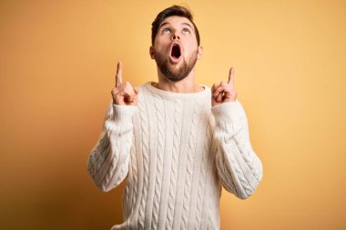 Sarışın, sakallı, mavi gözlü, sarı arka planda beyaz süveter giyen genç adam şaşırmış ve şaşırmış bir şekilde yukarı bakıyor ve parmaklarını gösterip kollarını kaldırıyordu..