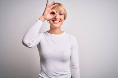 Genç, sarışın, modern kısa saçlı bir kadın izole edilmiş arka planda duruyor. El hareketleriyle gülümsüyor, gözleriyle mutlu yüzüyle parmaklarının arasından bakıyor..