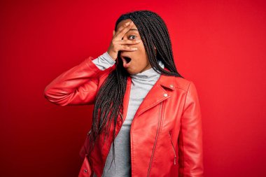 Genç Afro-Amerikan kadın, kırmızı izole edilmiş arka planda havalı deri ceket giyiyor. Şok olmuş bir şekilde yüzünü ve gözlerini kapıyor. Utanmış bir ifadeyle parmaklarının arasından bakıyor..