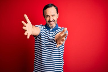 Orta yaşlı, beyaz, sıradan çizgili polo giyen, izole edilmiş kırmızı arka planda duran, kucaklaşmak için kollarını açıp gülümseyen kameraya bakan bir adam. Mutluluğu kucaklayan neşeli bir ifade..