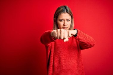 Güzel, sarışın bir kadın, kırmızı arka plan üzerine gündelik süveter giyiyor. Yumruğunu yumrukluyor, saldırıyor, saldırıyor, tehdit ediyor ve şiddet uyguluyor.