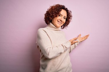 Orta yaşlı, kıvırcık saçlı, pembe arka plan üzerine balıkçı yaka kazak giyen güzel kadın gülümseyerek doğal bir şekilde içeri girmeye davet ediyor.