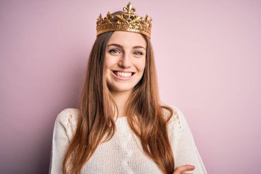 İzole edilmiş pembe arka plan üzerinde kraliçe tacı takan genç ve güzel bir kızıl kadın kollarını kavuşturup kameraya bakarken gülümsüyor. Pozitif kişi.