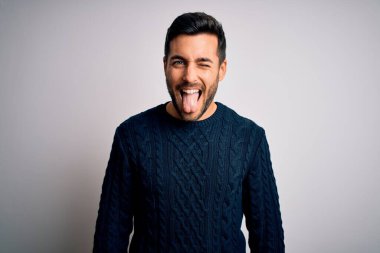 Sakallı, sıradan bir kazak giyen, beyaz arka planda dikilen, komik bir ifadeyle dilini dışarı çıkaran genç yakışıklı bir adam. Duygu kavramı.