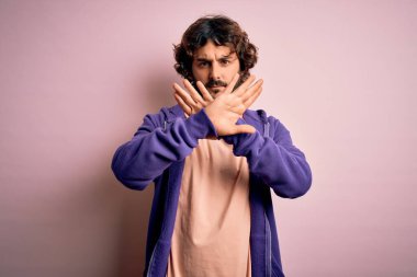 Genç, yakışıklı, sakallı, pembe arka planda sıradan bir kazak giyen, reddedilmiş, kollarını kavuşturup negatif işaret veren, sinirli yüzlü bir adam.