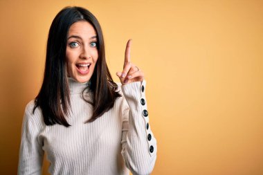 Mavi gözlü genç esmer kadın izole edilmiş sarı arka planda günlük süveter giyiyor ve başarılı bir fikirle işaret ediyor. Heyecanlı ve mutlu. Bir numara..