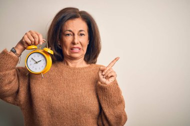 Orta yaşlı esmer kadın, izole edilmiş arka planda klasik alarm saatini tutuyor. Endişeli ve endişeli bir ifadeye işaret ediyor.