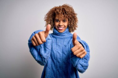 Beyaz arka plan üzerine balıkçı yaka kazak giyen genç ve güzel Afro-Amerikan kadın elleriyle olumlu jest yapmayı onaylıyor, gülümsüyor ve başarı için mutlu. Kazanan jest.