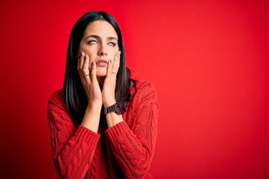 Mavi gözlü genç esmer kadın izole edilmiş kırmızı arka plan üzerine günlük süveter giyiyor. Yüzü, depresyonu ve üzüntüyü örten eller, sorun yüzünden üzgün ve tedirgin.