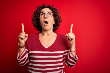 Orta yaşlı güzel kıvırcık saçlı kadın kırmızı arka planda sıradan çizgili süveter giyiyor şaşırmış ve şaşırmış bir şekilde yukarı bakıyor ve parmaklarıyla işaret ediyor ve kollarını kaldırıyor..