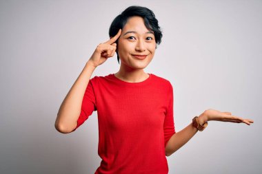 İzole edilmiş beyaz arka plan üzerinde duran sıradan kırmızı tişört giyen genç ve güzel Asyalı kız avuç içi açık fotokopi mekanı göstermekten ve alnını işaret etmekten rahatsız oluyor. Bunu bir düşün..