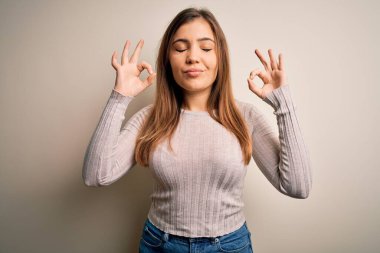 Güzel, genç bir kadın izole edilmiş arka planda rahat duruyor. Gözleri kapalı gülümsüyor ve parmaklarıyla meditasyon hareketi yapıyor. Yoga konsepti.