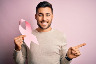 Elinde pembe kanser kurdelesi olan genç bir adam izole edilmiş arka planda desteğin sembolü çok mutlu bir şekilde elini ve parmağını kenarı işaret ediyor.
