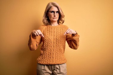 Orta yaşlı güzel sarışın kadın sarı arka planda rahat süveter ve gözlük takıyor üzgün ve üzgün görünüyor, parmaklarıyla yön gösteriyor, mutsuz ve depresif..