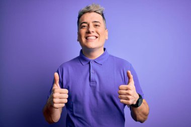İzole edilmiş arka plan başarısı tabelasının üzerine gündelik mor tişört giyen genç ve modern bir adam el hareketleriyle olumlu jest yapıyor, gülümsüyor ve mutlu. Neşeli ifade ve kazanan jest.