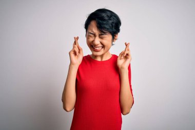 Genç, güzel Asyalı bir kız, günlük kırmızı tişört giyip izole edilmiş beyaz arka plan üzerinde duruyor. Umut ve gözler kapalı bir şekilde gülümseyerek işaret ediyor. Şans ve batıl inanç..