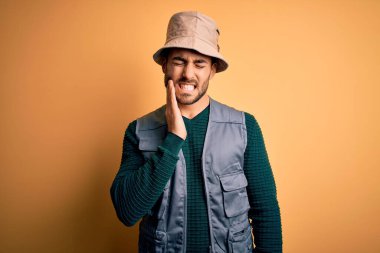 Tatilde sakallı yakışıklı turist sarı arka planda kaşif şapkası takıyor diş ağrısı ya da diş ağrısı yüzünden ağzıyla dokunuyor. Dişçi