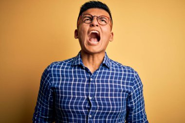 Genç, yakışıklı, latin gömlekli, sarı arka planda gözlüklü sinirli sinirli ve öfkeli bağıran öfkeyle bağıran bir adam. Öfke ve agresif kavram.