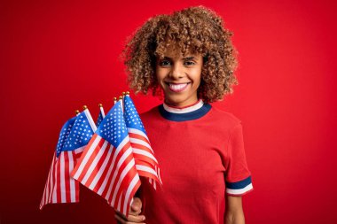 Genç Afro-Amerikan vatansever kıvırcık kadın kırmızı arka planda bayrakları tutuyor. Mutlu bir yüzü var ve gülümsüyor. Kendine gülerek dişlerini gösteriyor.