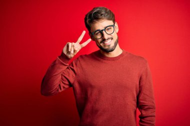 Sakallı, gözlüklü ve süveterli genç adam kırmızı arka planda duruyor. Barış sembolü yapıyor. Yüzünde parmaklar, gülümseyerek zaferini gösteriyor.