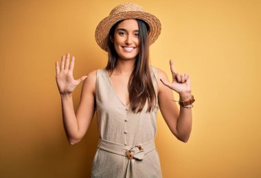 Genç, güzel, esmer kadın tatilde. Günlük elbiseler ve şapkalar giyiyor. Yedi numaralı parmaklarıyla işaret ediyor ve gülümsüyor..