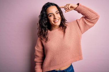Kıvırcık saçlı, rahat bir süveter giyen, pembe arka planda gülümseyen tek parmağıyla, harika bir fikir ya da düşünceyle kafasını gösteren güzel bir kadın.