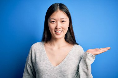 Genç ve güzel Asyalı bir kadın günlük süveter giyiyor mavi izole edilmiş arka planda duruyor neşeli bir şekilde gülümsüyor ve avuç içiyle kameraya bakıyor..