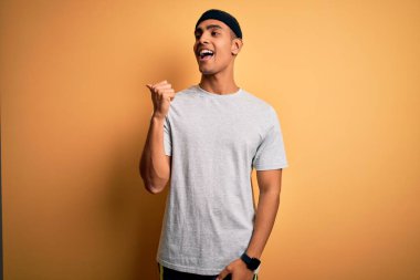 Yakışıklı Afro-Amerikan sporcusu, sarı arka planda spor giysisi giyiyor, gülümsüyor ve başparmağıyla yan tarafı işaret ediyor..